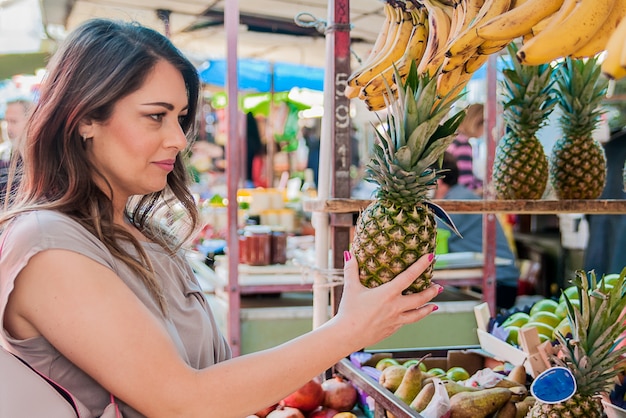 Atractiva mujer de compras en el mercado verde. Closeup retrato joven y bella mujer recogiendo, la elección de frutas, piñas. Expresión de la cara positiva emoción sensación de estilo de vida saludable