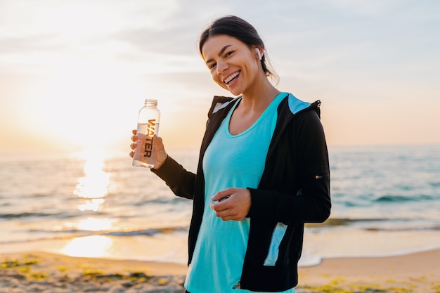 Atractiva mujer delgada haciendo ejercicios deportivos en la playa del amanecer de la mañana en ropa deportiva, agua potable sedienta en botella, estilo de vida saludable, escuchando música en auriculares inalámbricos, sonriendo feliz