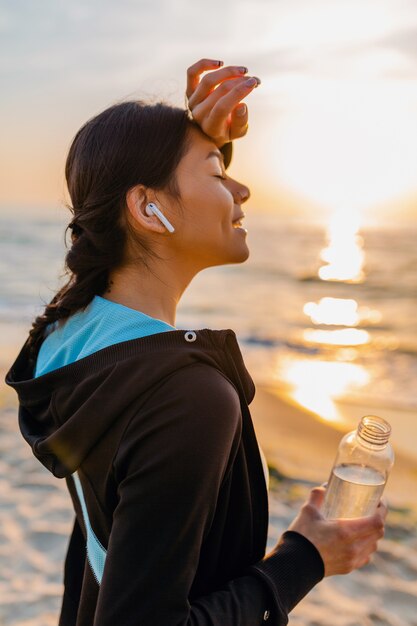 Atractiva mujer delgada haciendo ejercicios deportivos en la playa del amanecer de la mañana en ropa deportiva, agua potable sedienta en botella, estilo de vida saludable, escuchando música en auriculares inalámbricos, sonriendo feliz