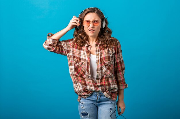 Atractiva mujer divertida con expresión de la cara sospechosa sorprendida mirando a un lado escuchando música en auriculares en camisa a cuadros y jeans aislados sobre fondo azul de estudio