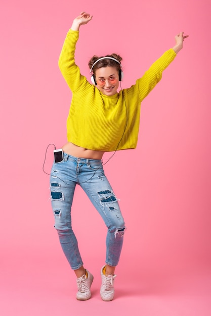 Foto gratuita atractiva mujer divertida feliz bailando escuchando música en auriculares vestida con traje de estilo colorido hipster aislado en pared rosa, vistiendo suéter amarillo y gafas de sol, divirtiéndose