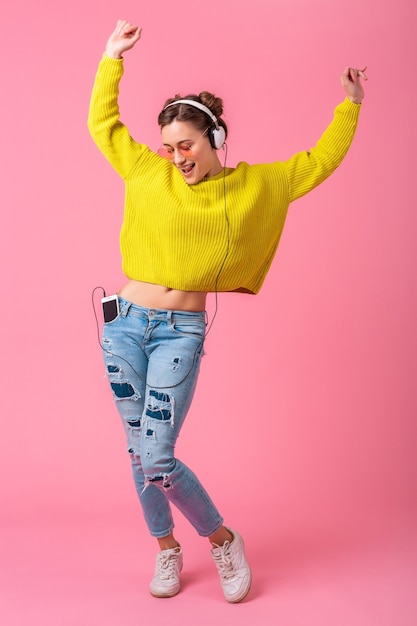 Foto gratuita atractiva mujer divertida feliz bailando escuchando música en auriculares vestida con traje de estilo colorido hipster aislado sobre fondo rosa studio