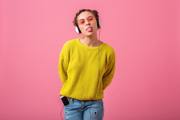 Atractiva mujer divertida feliz escuchando música en auriculares vestida con traje de estilo colorido hipster aislado en pared rosa, vistiendo suéter amarillo y gafas de sol, divirtiéndose