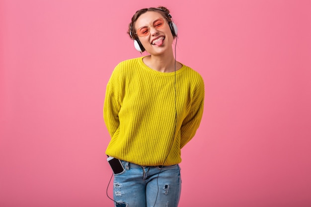 Atractiva mujer divertida feliz escuchando música en auriculares vestida con traje de estilo colorido hipster aislado en pared rosa, vistiendo suéter amarillo y gafas de sol, divirtiéndose