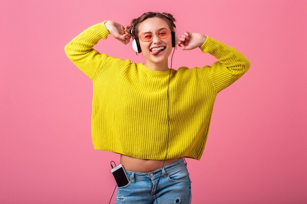 Atractiva mujer divertida feliz escuchando música en auriculares vestida con traje de estilo colorido hipster aislado en pared rosa, vistiendo suéter amarillo y gafas de sol, divirtiéndose