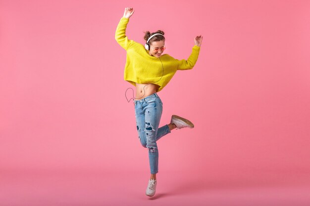 Atractiva mujer divertida feliz saltando escuchando música en auriculares vestida con traje de estilo colorido hipster aislado en pared rosa, vistiendo suéter amarillo y gafas de sol, divirtiéndose