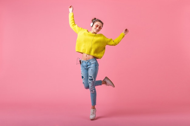 Atractiva mujer divertida feliz en suéter amarillo saltando escuchando música en auriculares vestida con traje de estilo colorido hipster aislado en la pared rosa, divirtiéndose