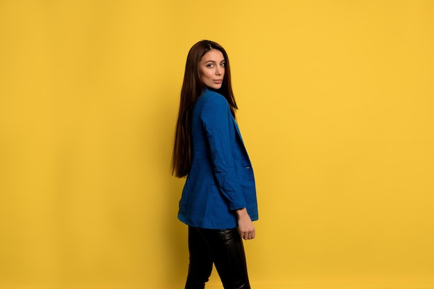 Atractiva mujer elegante con cabello largo oscuro con chaqueta azul posando sobre una pared aislada con una sonrisa encantadora