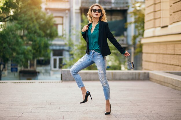 Atractiva mujer elegante caminando en la calle de la ciudad en zapatos de tacón, wesaring blue jeans, chaqueta negra, blusa verde, gafas de sol, sosteniendo un pequeño bolso, tendencia de moda de verano, delgada hermosa dama