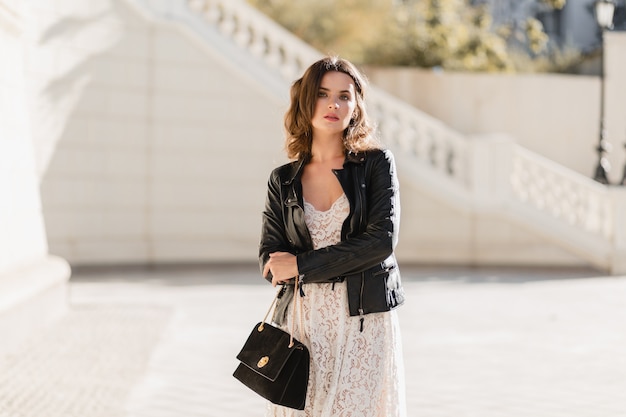 Atractiva mujer elegante caminando en la calle en traje de moda, sosteniendo el bolso, vestida con chaqueta de cuero negro y vestido de encaje blanco, estilo primavera otoño
