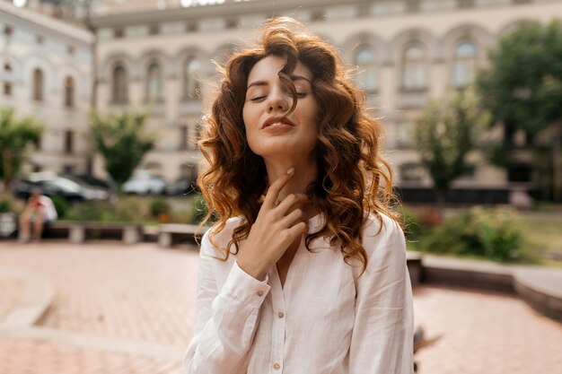 Atractiva mujer elegante con pelo rojo rizado vestido con camisa blanca tocando su cuello con los ojos cerrados y sonriendo