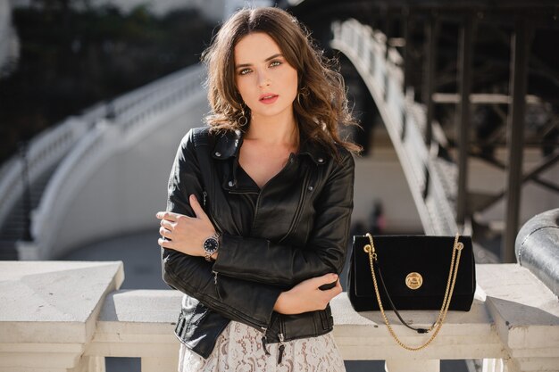 Atractiva mujer elegante posando en la calle en traje de moda, bolso de gamuza, con chaqueta de cuero negro y vestido de encaje blanco, estilo primavera otoño