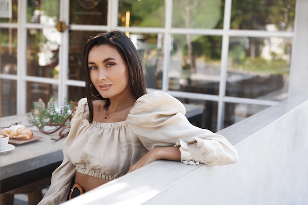 Atractiva mujer elegante sentada en la mesa de café al aire libre comiendo croissant y bebiendo café Modelo femenino sexy mira la cámara apoyada en la valla mientras disfruta de la comida en el restaurante cafetería