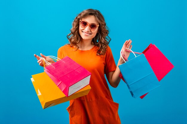 Atractiva mujer elegante sonriente emocional feliz adicta a las compras en vestido naranja de gran tamaño de moda con bolsas de la compra sobre fondo azul de estudio aislado