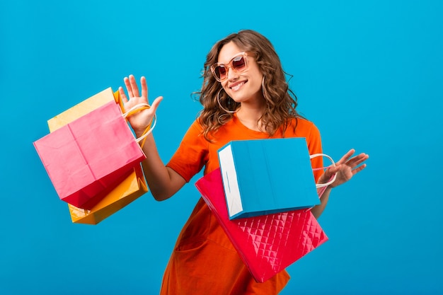 Atractiva mujer elegante sonriente emocional feliz adicta a las compras en vestido naranja de gran tamaño de moda con bolsas de la compra sobre fondo azul de estudio aislado