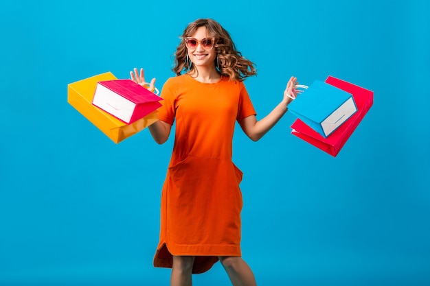 Atractiva mujer elegante sonriente feliz adicta a las compras en vestido naranja de gran tamaño de moda saltando corriendo sosteniendo bolsas de compras sobre fondo azul aislado