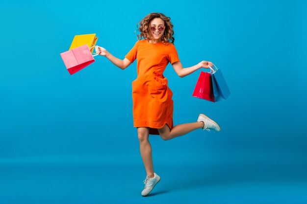 Atractiva mujer elegante sonriente feliz adicta a las compras en vestido naranja de gran tamaño de moda saltando sosteniendo bolsas de compras sobre fondo azul de estudio aislado