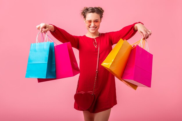 Atractiva mujer elegante sonriente feliz adicta a las compras en vestido rojo de moda con bolsas de colores en la pared rosa aislada, venta emocionada, tendencia de moda