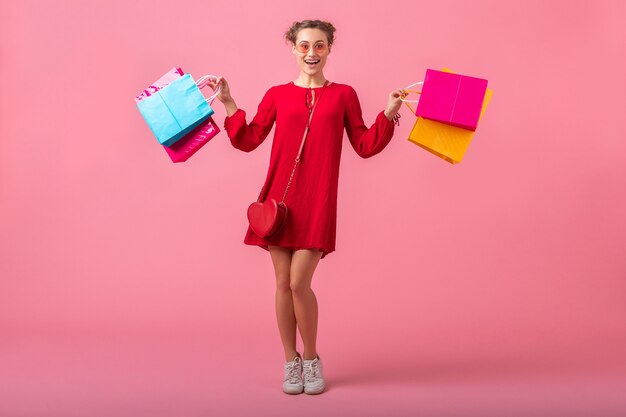 Atractiva mujer elegante sonriente feliz adicta a las compras en vestido rojo de moda con coloridas bolsas de compras en la pared rosa aislada, venta emocionada, tendencia de moda primavera verano