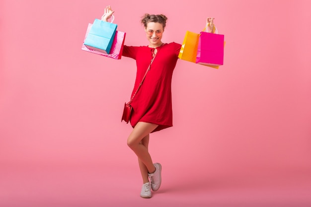 Atractiva mujer elegante sonriente feliz adicta a las compras en vestido rojo de moda con coloridas bolsas de compras en la pared rosa aislada, venta emocionada, tendencia de moda primavera verano