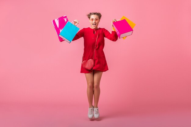 Atractiva mujer elegante sonriente feliz adicta a las compras en vestido rojo de moda con coloridas bolsas de compras en la pared rosa aislada, venta emocionada, tendencia de moda primavera verano