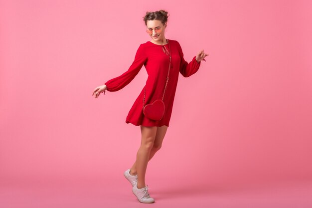 Atractiva mujer elegante sonriente feliz en vestido rojo de moda saltando bailando sobre pared rosa aislada, tendencia de moda primavera verano, chica coqueta de humor romántico