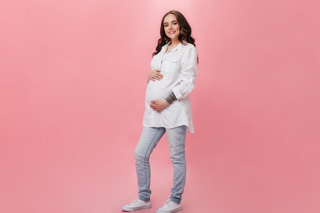 Atractiva mujer embarazada en camisa larga blanca sonríe ampliamente. feliz chica morena en jeans posa sobre fondo rosa.