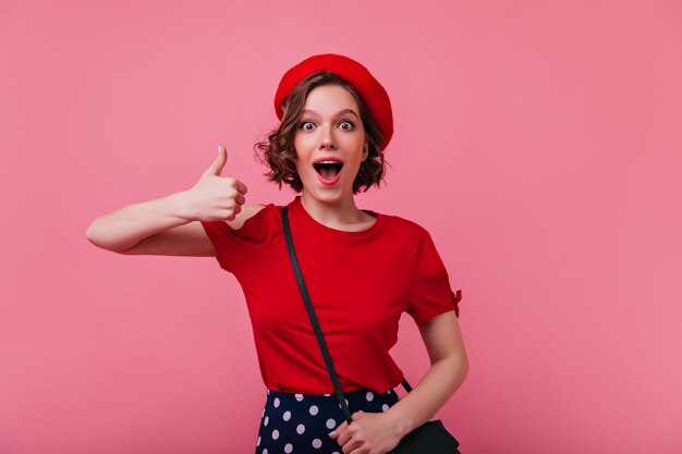 Atractiva mujer emocionada posando con el pulgar hacia arriba. Retrato de interior de alegre chica francesa en boina roja de moda.