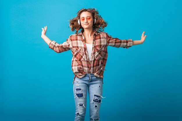 Atractiva mujer emocional sonriente saltando con expresión divertida cara loca en camisa a cuadros y jeans aislados sobre fondo azul de estudio, con gafas de sol rosa, salió mirando hacia arriba