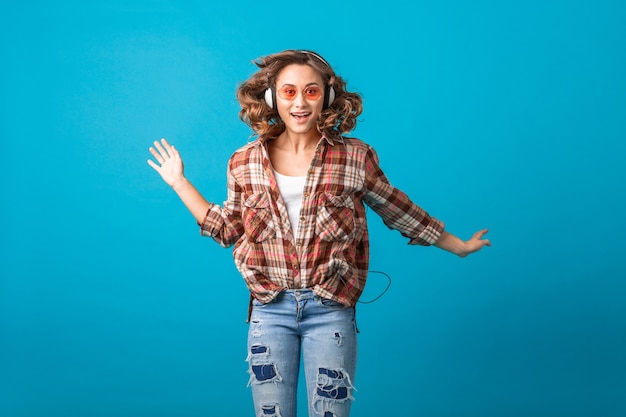 Atractiva mujer emocional sonriente saltando con expresión divertida cara loca en camisa a cuadros y jeans aislados sobre fondo azul de estudio, con gafas de sol rosa