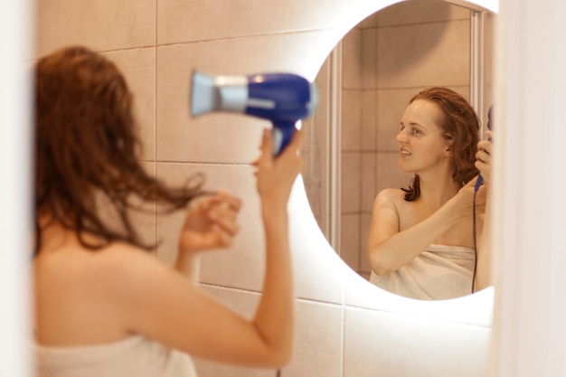 Atractiva mujer envuelta en una toalla blanca de pie con los hombros desnudos frente al espejo y secándose el cabello, haciendo procedimientos matutinos antes de ir a trabajar.