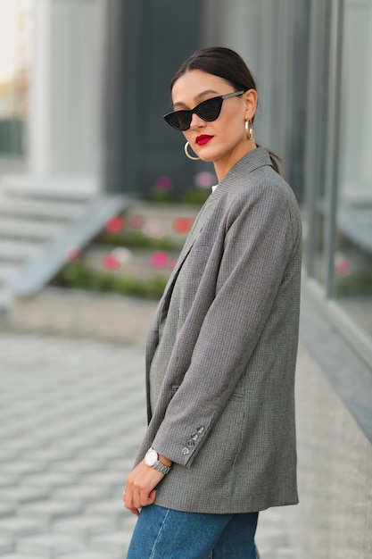 Atractiva mujer con estilo en gafas de sol con chaqueta gris posando con labios rojos al aire libre en la ciudad