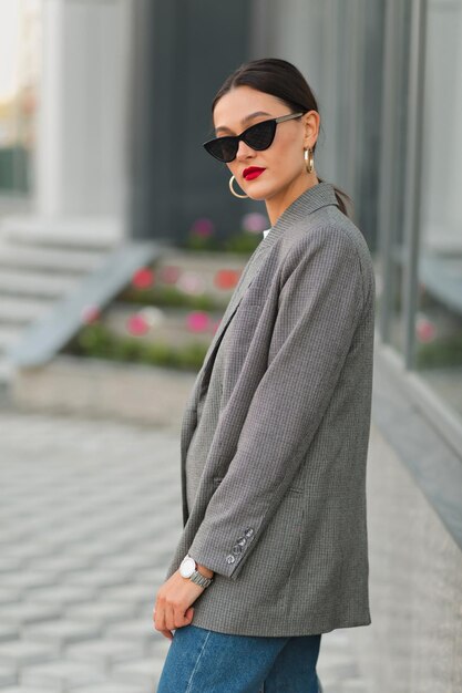 Atractiva mujer con estilo en gafas de sol con chaqueta gris posando con labios rojos al aire libre en la ciudad