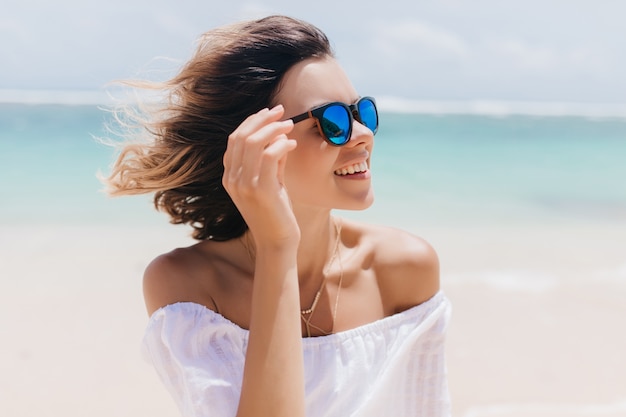 Atractiva mujer europea de pelo corto escalofriante en el resort. Increíble mujer bronceada en gafas de sol relajantes en la playa de arena en verano.