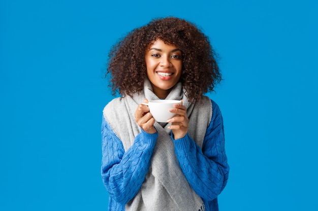 Atractiva mujer feliz afroamericana sonriente con pelo rizado, envuélvase con bufanda y tomando café, pared azul.