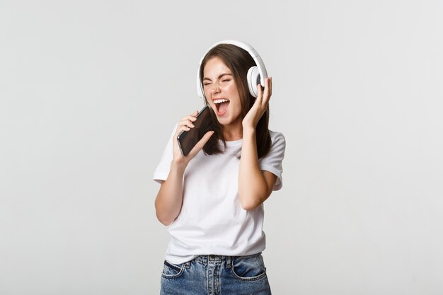 Atractiva mujer feliz jugando aplicación de karaoke, cantando en el teléfono inteligente, usando auriculares inalámbricos.