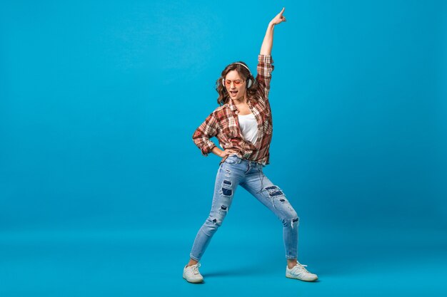 Atractiva mujer feliz posando de humor alegre escuchando música en auriculares con camisa a cuadros y jeans aislados sobre fondo azul de estudio, mirando hacia arriba