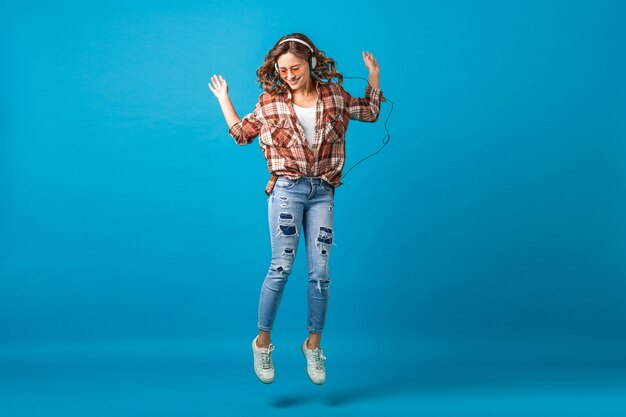 Atractiva mujer feliz saltando de humor alegre escuchando música en auriculares con camisa a cuadros y jeans aislado sobre fondo azul de estudio