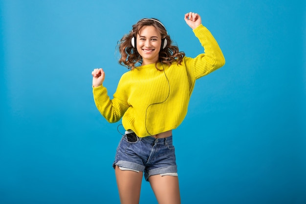 Atractiva mujer feliz sonriente bailando escuchando música en auriculares en traje elegante hipster aislado sobre fondo azul de estudio, vistiendo pantalones cortos y suéter amarillo