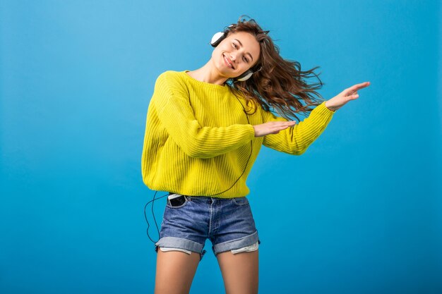 Atractiva mujer feliz sonriente bailando escuchando música en auriculares en traje elegante hipster aislado sobre fondo azul de estudio, vistiendo pantalones cortos y suéter amarillo