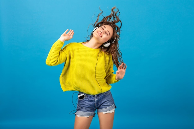 Atractiva mujer feliz sonriente bailando escuchando música en auriculares en traje elegante hipster aislado sobre fondo azul de estudio, vistiendo pantalones cortos y suéter amarillo