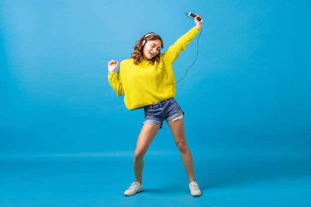 Atractiva mujer feliz sonriente bailando escuchando música en auriculares en traje hipster aislado sobre fondo azul de estudio, vistiendo pantalones cortos y suéter amarillo