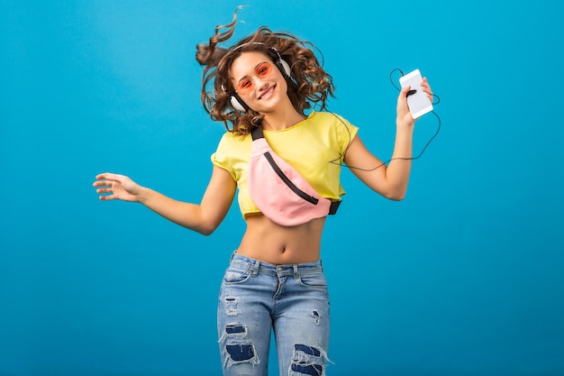 Foto gratuita atractiva mujer feliz sonriente bailando escuchando música en auriculares vestida con traje elegante hipster aislado sobre fondo azul de estudio, con ropa colorida