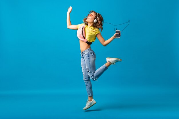 Atractiva mujer feliz sonriente bailando escuchando música en auriculares vestida con traje elegante hipster aislado sobre fondo azul de estudio, con ropa colorida