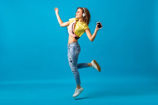 Atractiva mujer feliz sonriente bailando escuchando música en auriculares vestida con traje elegante hipster aislado sobre fondo azul de estudio, con ropa colorida