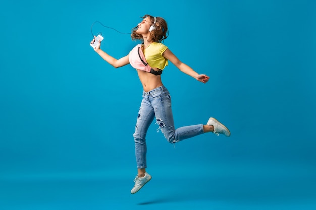 Atractiva mujer feliz sonriente bailando escuchando música en auriculares vestida con traje elegante hipster aislado sobre fondo azul de estudio, con ropa colorida