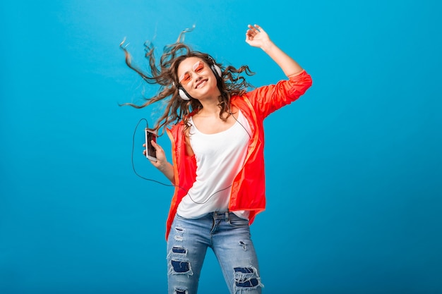 Atractiva mujer feliz sonriente bailando escuchando música en auriculares vestida con traje de estilo hipster aislado sobre fondo azul de estudio, con chaqueta rosa y gafas de sol