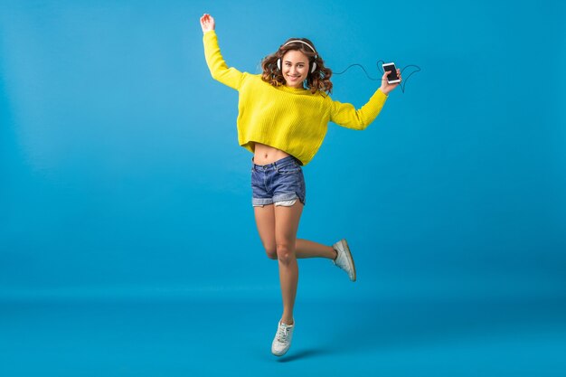 Atractiva mujer feliz sonriente saltando bailando escuchando música en auriculares en traje hipster aislado sobre fondo azul de estudio, vistiendo pantalones cortos y suéter amarillo