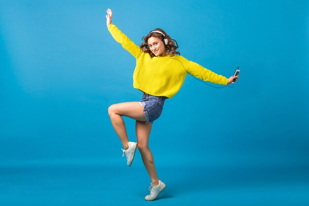 Atractiva mujer feliz sonriente saltando bailando escuchando música en auriculares en traje hipster aislado sobre fondo azul de estudio, vistiendo pantalones cortos y suéter amarillo