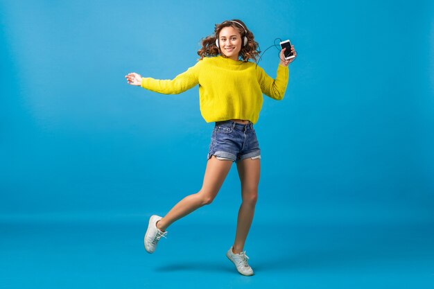 Atractiva mujer feliz sonriente saltando bailando escuchando música en auriculares en traje hipster aislado sobre fondo azul de estudio, vistiendo pantalones cortos y suéter amarillo